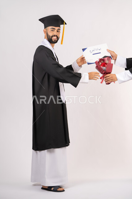 مفهوم التفوق والاجتهاد، استلام الشهادة بتعابير الفروح والسرور، بورتريه من الجانب لخريج عربي خليجي اماراتي يرتدي قبعة وثوب التخرج يقوم بأخذ باقة الورد بايماءات السعادة والسرور، الاحتفال بالنجاح وانهاء الدراسة الجامعية، صورة الجسم بالطول الكامل، خلفية بيضاء