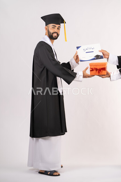 تقديم الهدايا في المناسبات السعيدة، استلام الشهادة بتعابير الفروح والسرور، مفهوم التفوق والاجتهاد، بورتريه من الجانب لخريج عربي خليجي اماراتي يرتدي قبعة وثوب التخرج يقوم بأخذ صندوق هدية،  الاحتفال بالنجاح وانهاء الدراسة الجامعية، الجسم بالطول الكامل، خلفية بيضاء