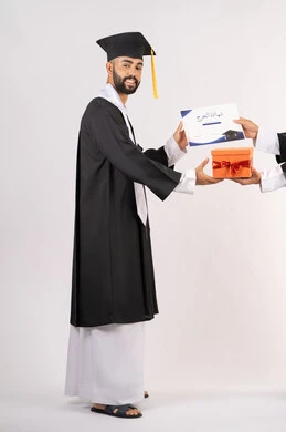 Giving gifts on happy occasions, receiving a certificate with expressions of joy and happiness, the concept of excellence and diligence, a side portrait of an Arab Gulf Emirati graduate wearing a graduation cap and gown taking a gift box to celebrate success and completion of university studies, full body, white background