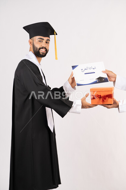التفوق والاجتهاد والنجاح، الاحتفال بانهاء الدراسة الجامعية، تقديم الهدايا في المناسبات السعيدة، استلام الشهادة بتعابير الفروح والسرور، بورتريه من الجانب لخريج عربي خليجي اماراتي يرتدي قبعة وثوب التخرج يقوم بأخذ صندوق هدية، خلفية بيضاء