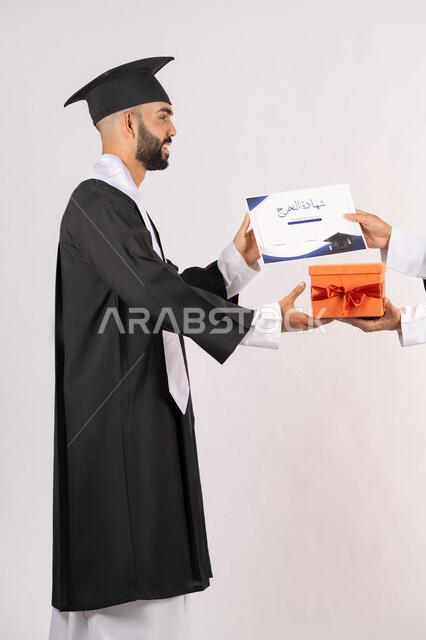 التفوق والاجتهاد والنجاح، الاحتفال بانهاء الدراسة الجامعية، تقديم الهدايا في المناسبات السعيدة، استلام الشهادة بتعابير الفروح والسرور، بورتريه من الجانب لخريج عربي خليجي اماراتي يرتدي قبعة وثوب التخرج يقوم بأخذ صندوق هدية، خلفية بيضاء