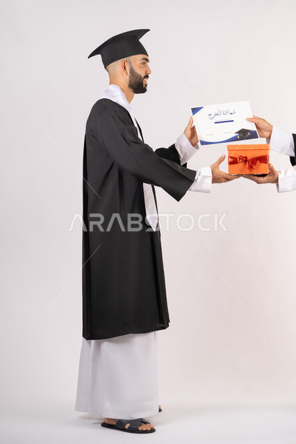 تقديم الهدايا في المناسبات السعيدة، استلام الشهادة بتعابير الفروح والسرور، مفهوم التفوق والاجتهاد، بورتريه من الجانب لخريج عربي خليجي اماراتي يرتدي قبعة وثوب التخرج يقوم بأخذ صندوق هدية،  الاحتفال بالنجاح وانهاء الدراسة الجامعية، الجسم بالطول الكامل، خلفية بيضاء
