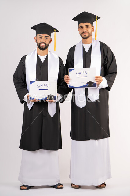 الاحتفال بإنهاء المرحلة الجامعية، الوقوف باستقامة والتقاط صور شخصية بثياب التخرج، بورتريه لشابين خريجين عربيين خليجيين اماراتيين مبتسمين يرتديان القبعة وعباءة التخرج ينظران الى الكاميرا بتعابير السرور، مفهوم التفوق والنجاح والاجتهاد، الجسم بالطول الكامل، خلفية بيضاء