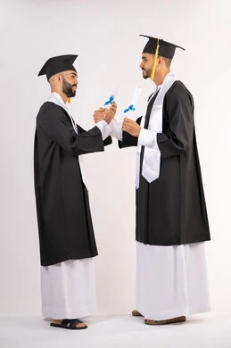 Taking selfies in graduation gowns, celebrating the completion of university, portrait of two smiling young Arab Gulf Emirati graduates wearing graduation cap and gown, standing straight with happy expressions, concept of excellence, success and diligence, full body, white background