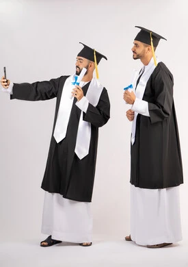 فرحة الحصول على شهادة التخرج، استخدام التقنية الحديثة، الاحتفال بإنهاء الدراسة الجامعية، الاحتفاظ بالذكريات السعيدة، بورتريه لشاب خريج عربي خليجي اماراتي يرتدي عباءه وقبعة التخرج يقوم بالتقاط صورة سيلفي تذكارية برفقة صديقه، الجسم بالطول الكامل، خلفية بيضاء