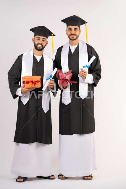 تقديم الهدايا في المناسبات السعيدة،  فرحة الحصول على شهادة التخرج، الاحتفال بإنهاء الدراسة الجامعية،  بورتريه لشاب خريج عربي خليجي اماراتي يرتدي عباءه وقبعة التخرج يقف مع صديقه ويمسكان بيديهما الشهادة مع باقة ورد مع صندوق هدية، الجسم بالطول الكامل، خلفية بيضاء