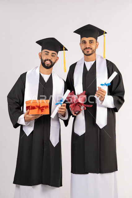 Obtaining a graduation certificate, giving gifts on happy occasions, celebrating the completion of university studies, a portrait of a young Arab Gulf Emirati graduate wearing an abaya and graduation cap standing with his friend, holding the certificate in their hands with a bouquet of flowers with a gift box, a university occasion and celebration, white background