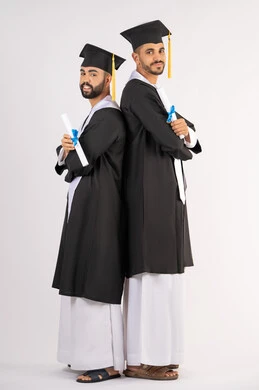 Celebrating the completion of university, standing up straight and taking selfies in graduation gowns, portrait of two smiling young Arab Gulf Emirati graduates wearing graduation cap and gown looking at the camera with happy expressions, concept of excellence, success and diligence, full body, white background