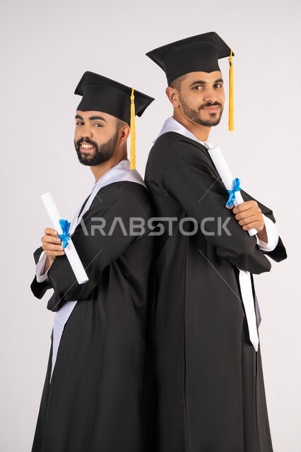 مناسبة واحتفال جامعي، الاحتفال بإنهاء المرحلة الجامعية، التقاط صور شخصية بثياب التخرج، بورتريه لشابين خريجين عربيين خليجيين اماراتيين مبتسمين يرتديان القبعة وعباءة التخرج ينظران الى الكاميرا بتعابير السرور، مفهوم التفوق والنجاح والاجتهاد، خلفية بيضاء