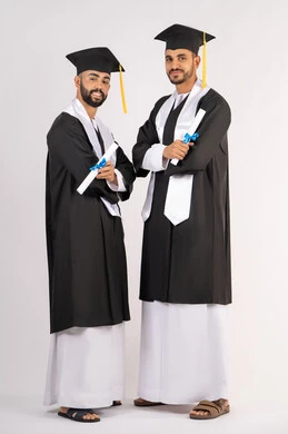 Celebrating the completion of university, standing up straight and taking selfies in graduation gowns, portrait of two smiling young Arab Gulf Emirati graduates wearing graduation cap and gown looking at the camera with happy expressions, concept of excellence, success and diligence, full body, white background