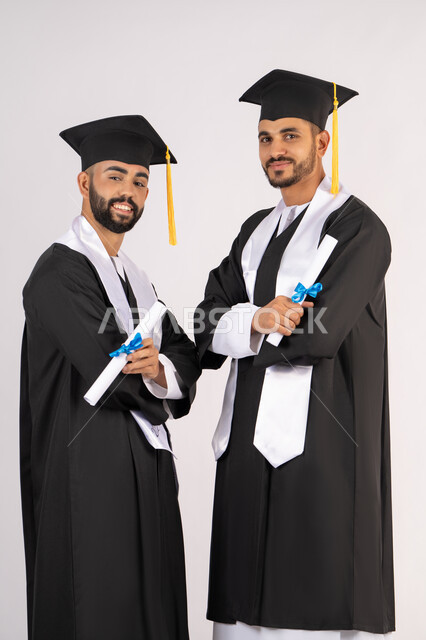 مناسبة واحتفال جامعي، الاحتفال بإنهاء المرحلة الجامعية، التقاط صور شخصية بثياب التخرج، بورتريه لشابين خريجين عربيين خليجيين اماراتيين مبتسمين يرتديان القبعة وعباءة التخرج ينظران الى الكاميرا بتعابير السرور، مفهوم التفوق والنجاح والاجتهاد، خلفية بيضاء