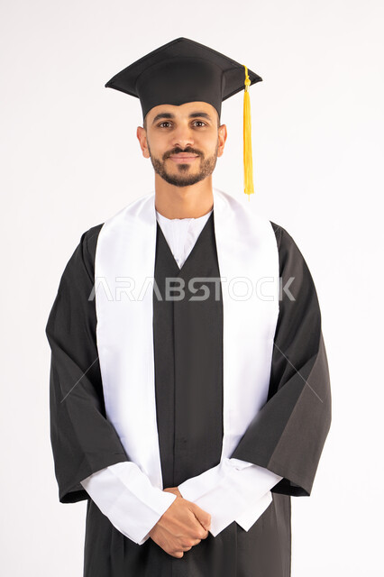 مفهوم التفوق والنجاح والاجتهاد، الاحتفال بإنهاء المرحلة الجامعية، الوقوف باستقامة والتقاط صور شخصية بثياب التخرج، بورتريه لشاب خريج عربي خليجي اماراتي مبتسم يرتدي القبعة وعباءة التخرج يتنظر الى الكاميرا بتعابير السرور، خلفية بيضاء
