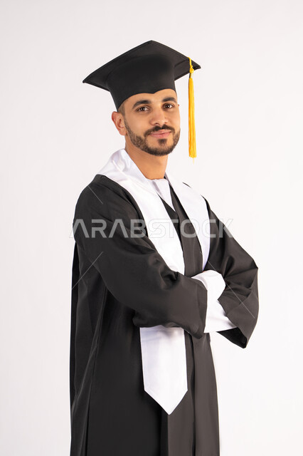 التقاط صور شخصية بثياب التخرج، الاحتفال بإنهاء المرحلة الجامعية، بورتريه لشاب خريج عربي خليجي اماراتي مبتسم يرتدي القبعة وعباءة التخرج يقف بأيدي متقاطعة ينظر الى الكاميرا بتعابير الثقة بالنفس، مفهوم التفوق والنجاح والاجتهاد، خلفية بيضاء