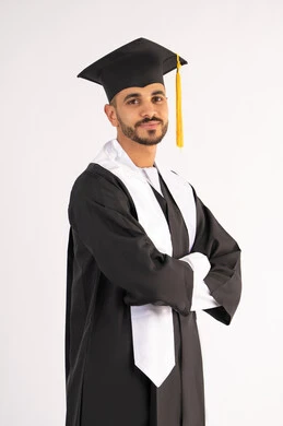 Taking selfies in graduation gowns, celebrating the completion of university, portrait of a smiling young Arab Gulf Emirati graduate wearing a graduation cap and gown, standing with crossed hands looking at the camera with an expression of self-confidence, the concept of excellence, success and diligence, white background