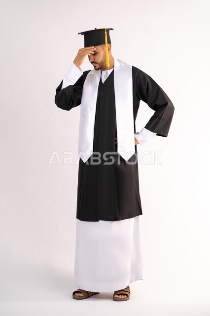 Waiting impatiently for results, exhaustion and fatigue after hard work and perseverance, finishing university studies after suffering, portrait of a young Arab Gulf Emirati graduate wearing a cap and graduation gown, putting his hand on his head with an expression of despair, full body, white background