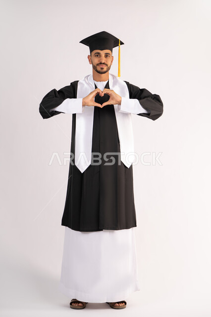 تشكيل اليدين على شكل قلب، التعبير عن الحب والفرح، التقاط صور شخصية بثياب التخرج، الاحتفال بإنهاء المرحلة الجامعية بنجاح وتفوق، بورتريه لشاب خريج عربي خليجي اماراتي مبتسم يرتدي القبعة وعباءة التخرج ينظر الى الكاميرا بتعابير السرور، الجسم بالطول الكامل، خلفية بيضاء