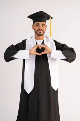 تشكيل اليدين على شكل قلب، التعبير عن الحب والفرح، التقاط صور شخصية بثياب التخرج، الاحتفال بإنهاء المرحلة الجامعية بنجاح وتفوق، بورتريه لشاب خريج عربي خليجي اماراتي مبتسم يرتدي القبعة وعباءة التخرج ينظر الى الكاميرا بتعابير السرور، خلفية بيضاء