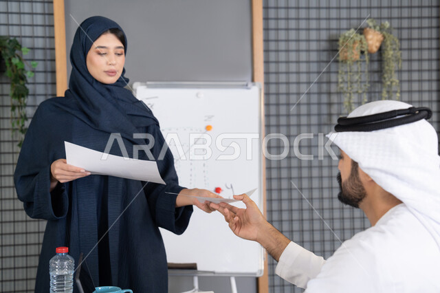 إدارة و تقسيم المهام، إجراء اجتماع عمل داخل غرفة الاجتماعات في مقر الشركة، شرح و تحليل الرسوم البيانية، إمرأة عربية خليجية اماراتية ترتدي الحجاب والعباءة تقوم بتوزيع اوراق النتائج على زملائها، دراسة أوراق البيانات الإحصائية، تطوير المشاريع و الاستثمارات