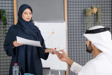 إدارة و تقسيم المهام، إجراء اجتماع عمل داخل غرفة الاجتماعات في مقر الشركة، شرح و تحليل الرسوم البيانية، إمرأة عربية خليجية اماراتية ترتدي الحجاب والعباءة تقوم بتوزيع اوراق النتائج على زملائها، دراسة أوراق البيانات الإحصائية، تطوير المشاريع و الاستثمارات