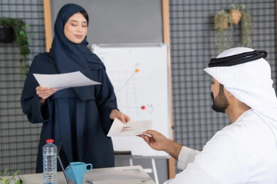 دراسة أوراق البيانات الإحصائية, تطوير المشاريع و الاستثمارات, إدارة و تقسيم المهام، إجراء اجتماع عمل داخل غرفة الاجتماعات في مقر الشركة، شرح و تحليل الرسوم البيانية، إمرأة عربية خليجية اماراتية ترتدي الحجاب والعباءة تقوم بتوزيع اوراق النتائج على زملائها