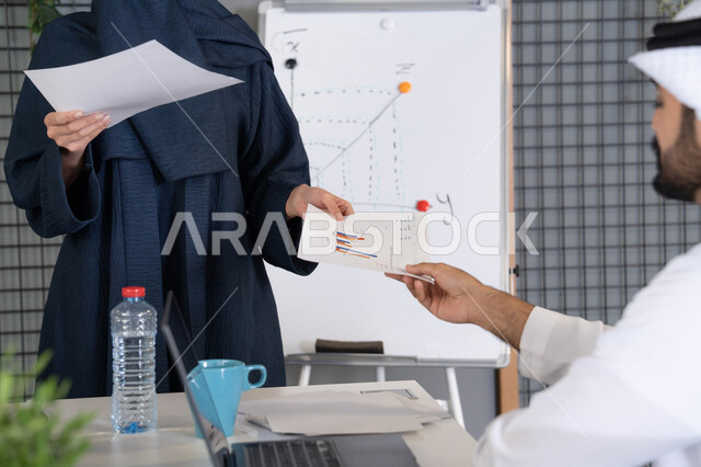 Project and investment development, managing and dividing tasks, conducting a business meeting in the company's headquarters meeting room, explaining and analyzing graphs, an Emirati Gulf Arab woman wearing a hijab and abaya distributing results sheets to her colleagues, studying statistical data sheets