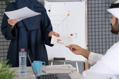 تطوير المشاريع و الاستثمارات, إدارة و تقسيم المهام، إجراء اجتماع عمل داخل غرفة الاجتماعات في مقر الشركة، شرح و تحليل الرسوم البيانية، إمرأة عربية خليجية اماراتية ترتدي الحجاب والعباءة تقوم بتوزيع اوراق النتائج على زملائها، دراسة أوراق البيانات الإحصائية