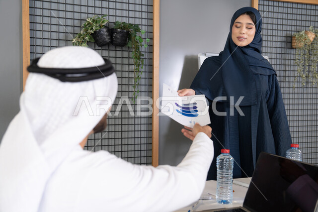 Managing and dividing tasks, conducting a business meeting in the company's headquarters meeting room, explaining and analyzing graphs, an Emirati Gulf Arab woman wearing a hijab and abaya distributing results sheets to her colleagues, studying statistical data sheets, developing projects and investments
