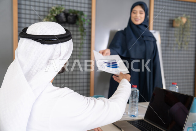 Project and investment development, managing and dividing tasks, conducting a business meeting in the company's headquarters meeting room, explaining and analyzing graphs, an Emirati Gulf Arab woman wearing a hijab and abaya distributing results sheets to her colleagues, studying statistical data sheets