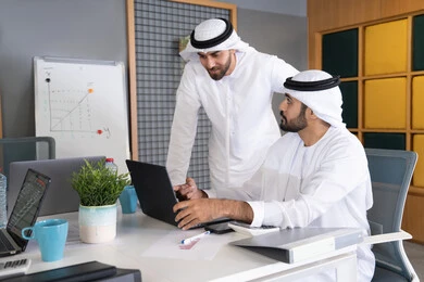 Following and watching business projects on a laptop, two Emirati Gulf Arab men wearing kandura and ghutra completing work tasks via computer, cooperation between employees to achieve company goals, integrating work with technology and technology, the development of Emirati companies