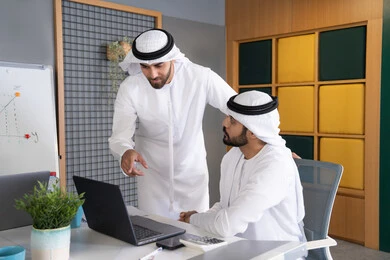 مشاهدة ومتابعة مشاريع الأعمال على الحاسب المحمول، رجلان خليجيان عربيان اماراتيان يرتديان الكندروة والغترة يقومان بإنجاز مهام العمل عبر جهاز الحاسوب، التعاون بين الموظفين لتحقيق أهداف الشركة، دمج العمل مع التقنية والتكنولوجيا، تطور الشركات الاماراتية