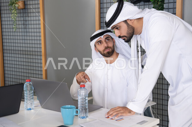 دمج العمل مع التقنية والتكنولوجيا، تطور الشركات الاماراتية، متابعة ومشاهدة مشاريع الأعمال على الحاسب المحمول، رجلان خليجيان عربيان اماراتيان يرتديان الكندروة والغترة يقومان بإنجاز مهام العمل عبر جهاز الحاسوب، التعاون بين الموظفين لتحقيق أهداف الشركة