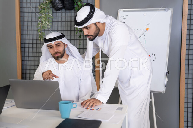 التعاون بين الموظفين لتحقيق أهداف الشركة، دمج العمل مع التقنية والتكنولوجيا، تطور الشركات الاماراتية، متابعة ومشاهدة مشاريع الأعمال على الحاسب المحمول، رجلان خليجيان عربيان اماراتيان يرتديان الكندروة والغترة يقومان بإنجاز مهام العمل عبر جهاز الحاسوب