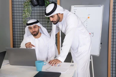 التعاون بين الموظفين لتحقيق أهداف الشركة، دمج العمل مع التقنية والتكنولوجيا، تطور الشركات الاماراتية، متابعة ومشاهدة مشاريع الأعمال على الحاسب المحمول، رجلان خليجيان عربيان اماراتيان يرتديان الكندروة والغترة يقومان بإنجاز مهام العمل عبر جهاز الحاسوب
