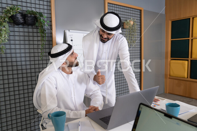 رجلان خليجيان عربيان اماراتيان يرتديان الكندروة والغترة يقومان بإنجاز مهام العمل عبر جهاز الحاسوب، التعاون بين الموظفين لتحقيق أهداف الشركة، دمج العمل مع التقنية والتكنولوجيا، تطور الشركات الاماراتية، متابعة ومشاهدة مشاريع الأعمال على الحاسب المحمول