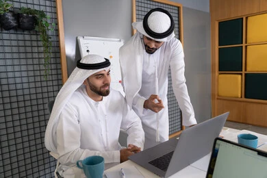 دمج العمل مع التقنية والتكنولوجيا، تطور الشركات الاماراتية، متابعة ومشاهدة مشاريع الأعمال على الحاسب المحمول، رجلان خليجيان عربيان اماراتيان يرتديان الكندروة والغترة يقومان بإنجاز مهام العمل عبر جهاز الحاسوب، التعاون بين الموظفين لتحقيق أهداف الشركة