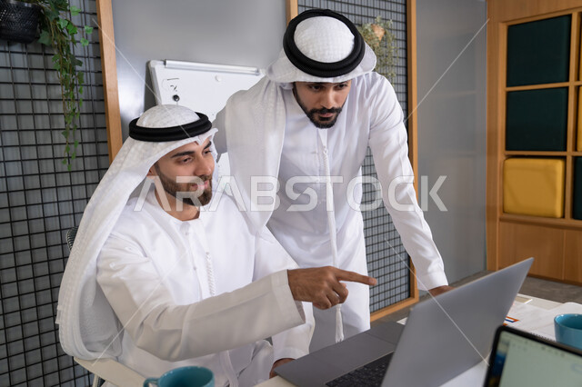 التعاون بين الموظفين لتحقيق أهداف الشركة، دمج العمل مع التقنية والتكنولوجيا، تطور الشركات الاماراتية، متابعة ومشاهدة مشاريع الأعمال على الحاسب المحمول، رجلان خليجيان عربيان اماراتيان يرتديان الكندروة والغترة يقومان بإنجاز مهام العمل عبر جهاز الحاسوب