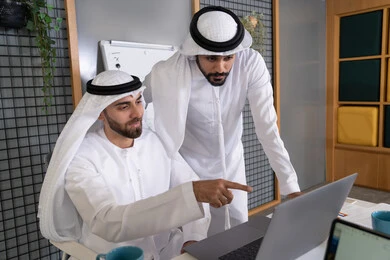 التعاون بين الموظفين لتحقيق أهداف الشركة، دمج العمل مع التقنية والتكنولوجيا، تطور الشركات الاماراتية، متابعة ومشاهدة مشاريع الأعمال على الحاسب المحمول، رجلان خليجيان عربيان اماراتيان يرتديان الكندروة والغترة يقومان بإنجاز مهام العمل عبر جهاز الحاسوب
