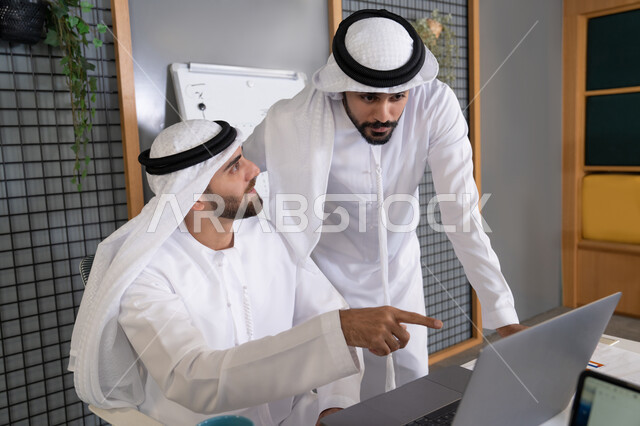 مشاهدة ومتابعة مشاريع الأعمال على الحاسب المحمول، رجلان خليجيان عربيان اماراتيان يرتديان الكندروة والغترة يقومان بإنجاز مهام العمل عبر جهاز الحاسوب، التعاون بين الموظفين لتحقيق أهداف الشركة، دمج العمل مع التقنية والتكنولوجيا، تطور الشركات الاماراتية