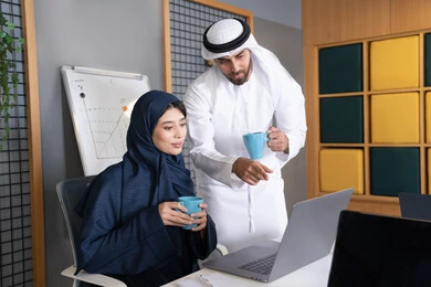 العمل الجماعي وروح الفريق الواحد، بيئة العمل المريحة في الامارات، تعليم الموظفين الأساليب الجديدة في العمل لتحقيق أهداف الشركة، دمج العمل مع التقنية والتكنولوجيا، زميلان عربيان خليجيان اماراتيان يقومان بإنجاز المهام عبر الحاسوب المحمول، الاشارة الى اللابتوب
