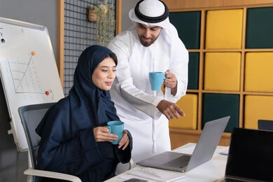 Training employees on new work methods to achieve company goals, integrating work with technology, two Emirati Gulf Arab colleagues completing tasks using laptops, pointing to the laptop, teamwork and team spirit, comfortable work environment in the UAE