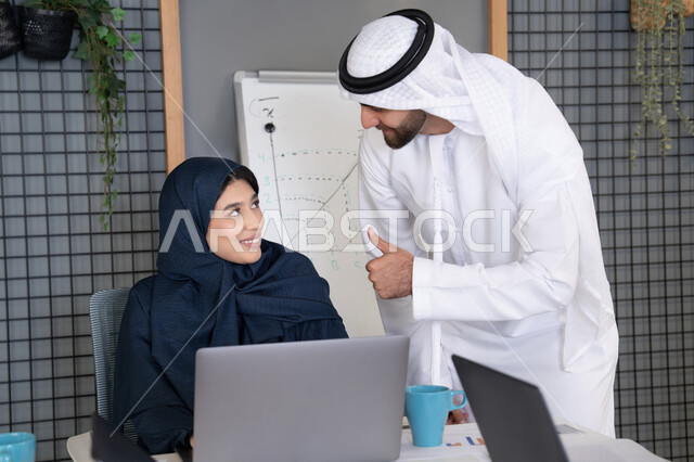 Teaching employees new work methods to achieve company goals, integrating work with technology, two Emirati Gulf Arab colleagues completing tasks using laptops, pointing to the laptop, teamwork and team spirit, comfortable work environment in the Emirates