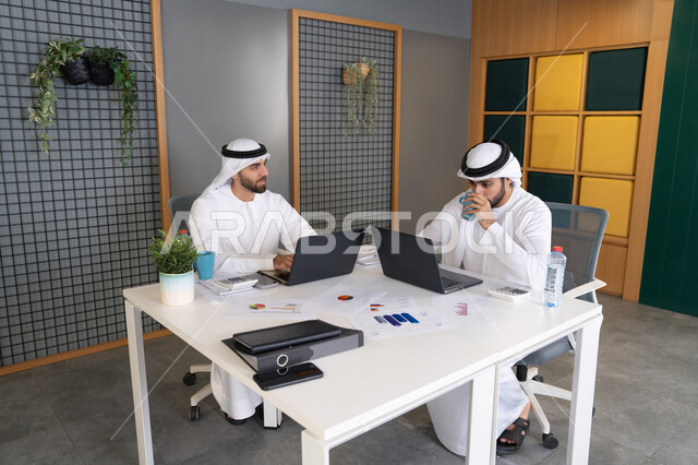 Integrating work with technology, the development of Emirati companies, following up on business projects, two Emirati Gulf Arab men wearing kandura and ghutra completing work tasks via computer, expressions and gestures of concentration, working on a plan to achieve the company's goals