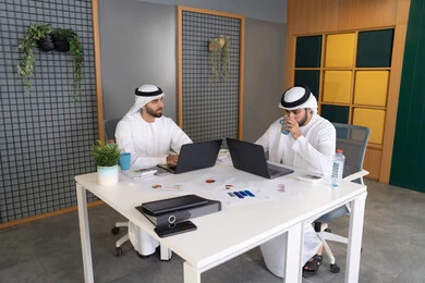 Integrating work with technology, the development of Emirati companies, following up on business projects, two Emirati Gulf Arab men wearing kandura and ghutra completing work tasks via computer, expressions and gestures of concentration, working on a plan to achieve the company's goals