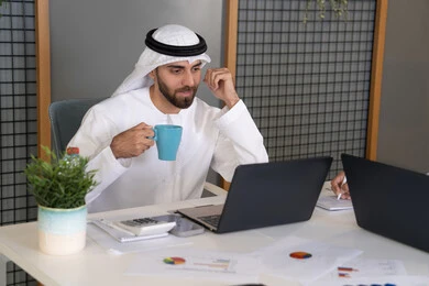 ايماءات وتعابير التركيز، العمل على الخطة لتحقيق اهداف الشركة، دمج العمل مع التقنية والتكنولوجيا، تطور الشركات الاماراتية، متابعة مشاريع الأعمال، رجل خليجي عربي اماراتي يرتدي الكندروة والغترة يقوم بإنجاز مهام العمل عبر جهاز الحاسوب