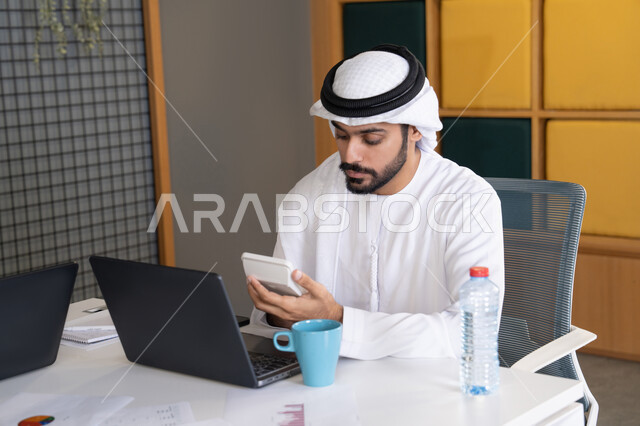 Gestures and expressions of concentration, working on a plan to achieve company goals, integrating work with technology, development of Emirati companies, following up on business projects, an Emirati Arab Gulf man wearing a kandura and ghutra completing work tasks via a computer