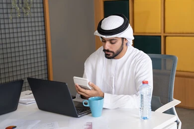 ايماءات وتعابير التركيز، العمل على الخطة لتحقيق اهداف الشركة، دمج العمل مع التقنية والتكنولوجيا، تطور الشركات الاماراتية، متابعة مشاريع الأعمال، رجل خليجي عربي اماراتي يرتدي الكندروة والغترة يقوم بإنجاز مهام العمل عبر جهاز الحاسوب