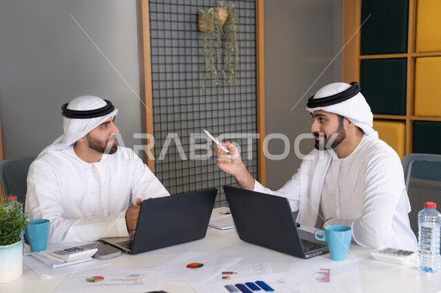 تطور الشركات الاماراتية، متابعة مشاريع الأعمال، رجلان خليجيان عربيان اماراتيان يرتديان الكندروة والغترة يقومان بإنجاز مهام العمل عبر جهاز الحاسوب، تعابير وايماءات التركيز، العمل على الخطة لتحقيق اهداف الشركة، دمج العمل مع التقنية والتكنولوجيا