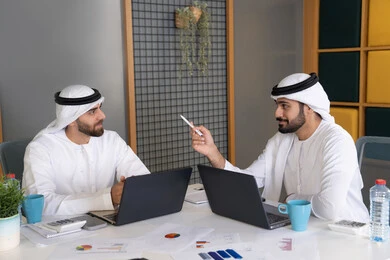 Development of Emirati companies, following up on business projects, two Emirati Gulf Arab men wearing kandura and ghutra completing work tasks via computer, expressions and gestures of concentration, working on a plan to achieve the company's goals, integrating work with technology and technology
