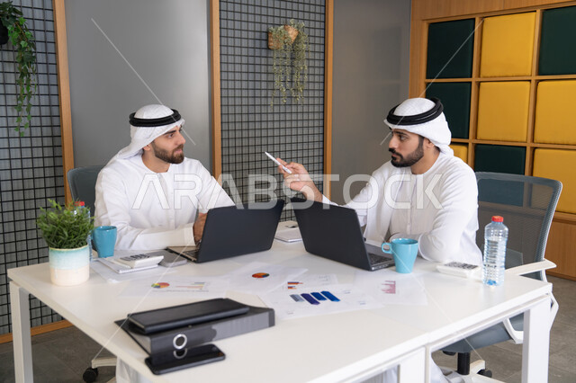 تعابير وايماءات التركيز، العمل على الخطة لتحقيق اهداف الشركة، دمج العمل مع التقنية والتكنولوجيا، تطور الشركات الاماراتية، متابعة مشاريع الأعمال، رجلان خليجيان عربيان اماراتيان يرتديان الكندروة والغترة يقومان بإنجاز مهام العمل عبر جهاز الحاسوب