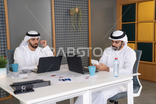 دمج العمل مع التقنية والتكنولوجيا، تطور الشركات الاماراتية، متابعة مشاريع الأعمال، رجلان خليجيان عربيان اماراتيان يرتديان الكندروة والغترة يقومان بإنجاز مهام العمل عبر جهاز الحاسوب، تعابير وايماءات التركيز، العمل على الخطة لتحقيق اهداف الشركة